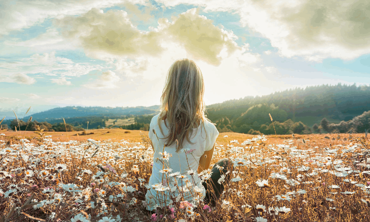 Choosing the Right Venue for a Funeral Service A woman sitting in a field of white flowers, looking out towards distant hills beneath a bright, cloudy sky.