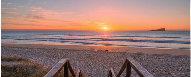 Sunrise over a sandy beach with calm ocean waves and a small island in the distance.